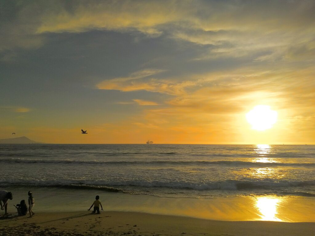 Sunset over Playa Hermosa beach in Ensenada, Baja California, with gentle waves and beachgoers.