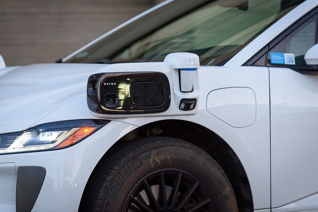 Close-up of a sensor package on the side of a white Waymo Jaguar I-PACE self-driving car, showing the camera and lidar units