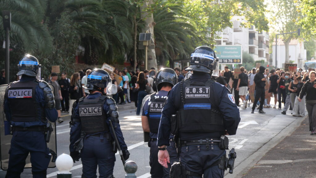 French riot police in helmets facing teenage student protesters holding signs during a high-school demonstration in Perpignan, France, October 2026.