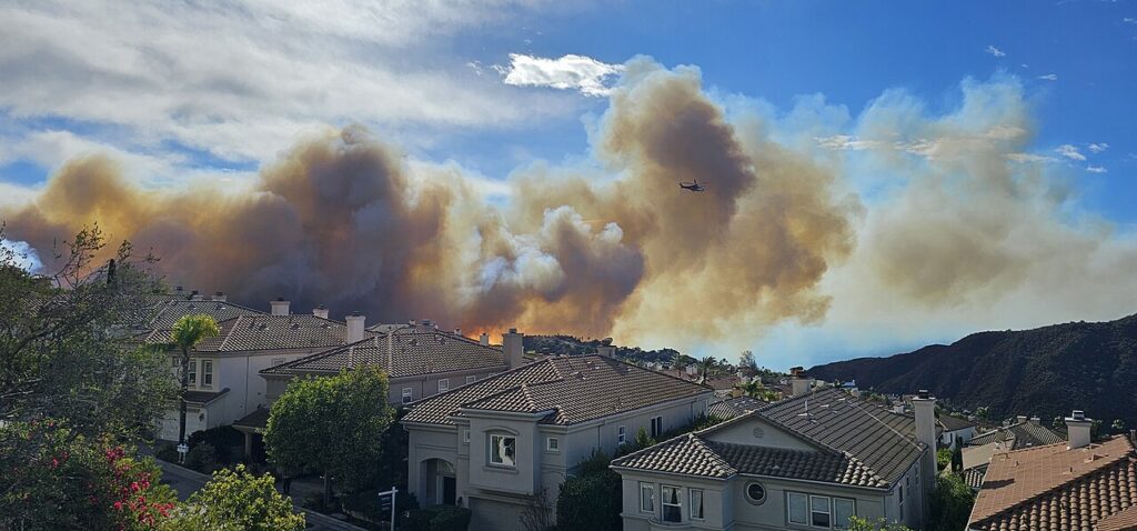 Wildfire flames and a towering smoke plume behind suburban homes in Southern California (file photo from the January 2025 Palisades Fire)