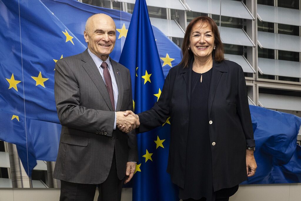 US ambassador to the EU Andrew Puzder shaking hands with European Commissioner Dubravka Suica in front of EU flags in Brussels.